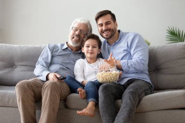 Neşeli büyükbaba yetişkin oğul ve torun TV seyredip patlamış mısır yiyor.