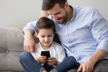 Baba ve küçük oğlu akıllı telefon kullanarak kanepede yan yana oturuyor.