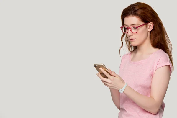 Young concentrated red-haired girl in eyewear holding smartphone. — ストック写真