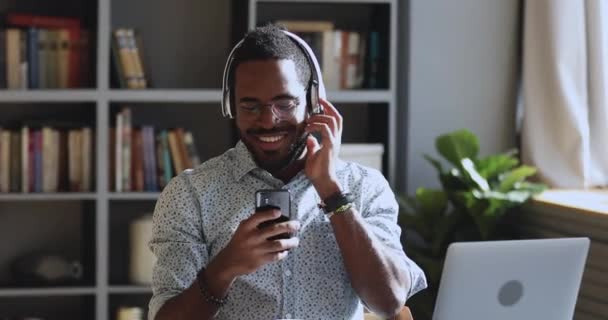 戴着耳机笑着的非洲男人在办公桌前听移动音乐 — 图库视频影像