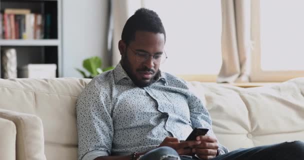 Sérieux jeune homme africain en utilisant l'application smartphone assis sur le canapé — Video