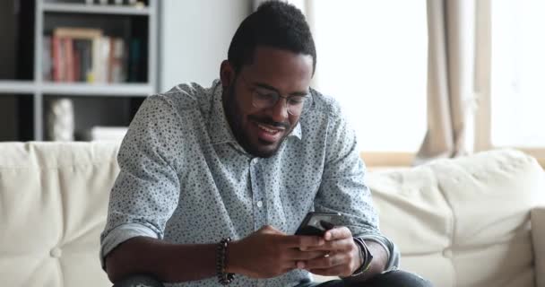 Sonriente chico africano usando el teléfono inteligente sentado en el sofá — Vídeo de stock