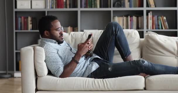 Young african guy using phone relax on sofa at home — Stock Video