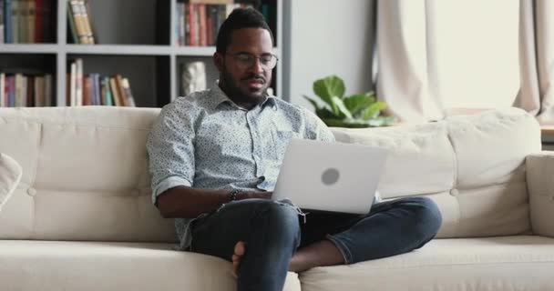 Allvarligt ung afrikansk man användare som håller laptop sitta på soffan — Stockvideo
