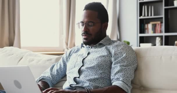 在家里工作的专注的非洲男人在沙发上使用笔记本电脑 — 图库视频影像