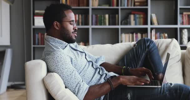Avslappnad allvarlig afrikansk kille som använder laptop på soffan hemma — Stockvideo