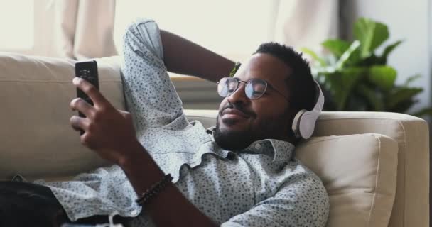 Afrikaanse man draagt koptelefoon ontspannen op de bank luisteren mobiele muziek — Stockvideo