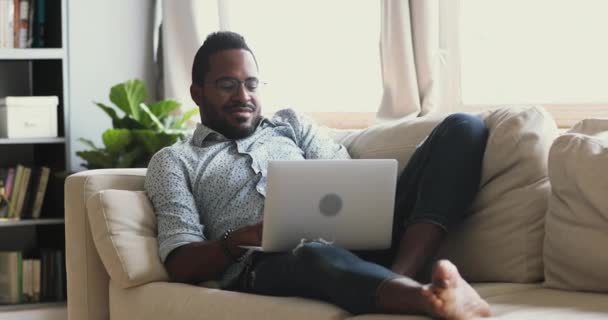 Mutlu Afrikalı adam kanepeye oturup dizüstü bilgisayar kullanarak hayatın tadını çıkarıyor. — Stok video