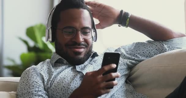 幸せなアフリカ人男身に着けていますヘッドフォンリスニングモバイル音楽上のソファ — ストック動画