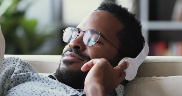 Безмятежный африканский мужчина носит наушники слушать музыку медитировать на диване — стоковое видео