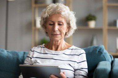 Odaklanmış yaşlı bir kadın elinde tablet bilgisayarla koltukta oturuyor.
