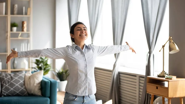 Excited independent indian woman stand in living room arms outstretched