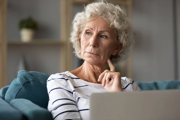 Pensive femme âgée assis sur le canapé avec ordinateur portable détourner les yeux — Photo