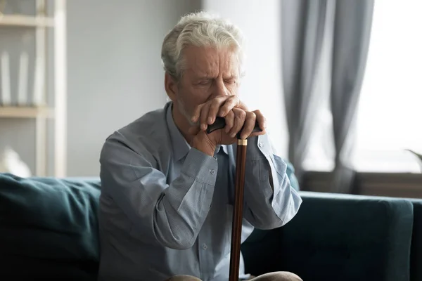 Depressed disabled retired man sitting on couch with cane stick — Stock Photo, Image