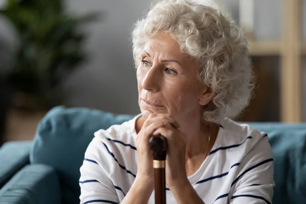 Disabled mature woman sit on sofa holding cane looking away