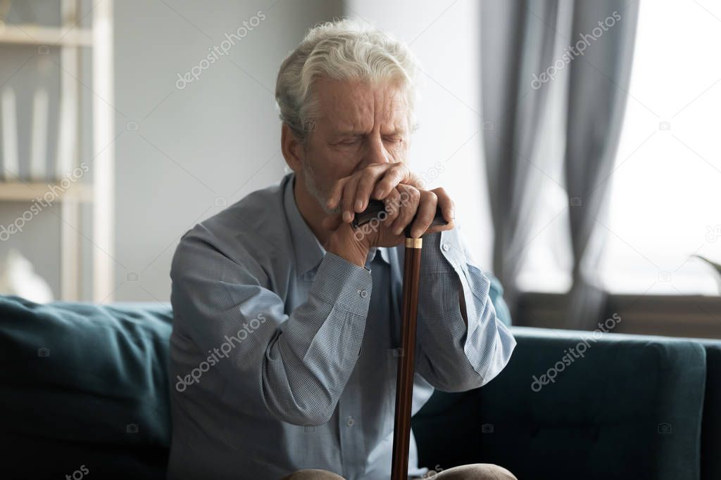 Depressed disabled retired man sitting on couch with cane stick