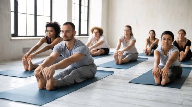 Eğitmenle yoga yapan farklı insanlar, eğilip eğilme egzersizi yapıyorlar.