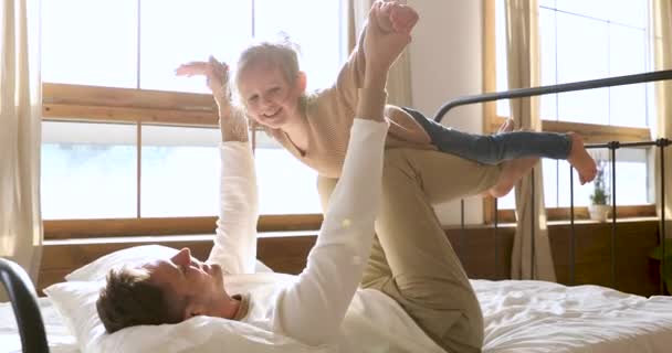 Маленька дочка летить як літак, граючи з батьком у ліжку — стокове відео