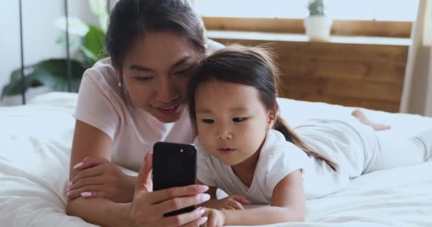 幸せなアジアの母親使用スマートフォンとともに子供の娘でベッド — ストック動画