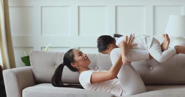 Feliz ásia mamãe jogar levantamento pequeno criança filha no sofá — Vídeo de Stock