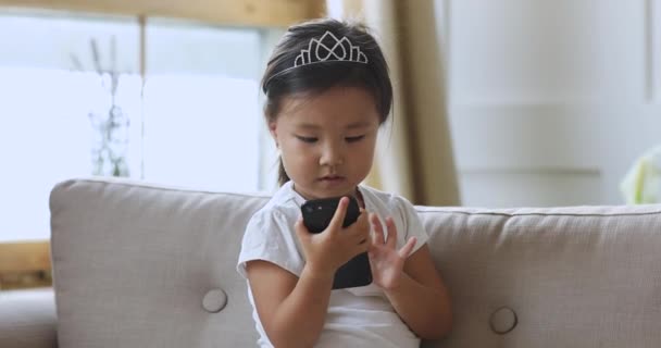 好奇心旺盛な小アジアの女の子一人でソファに座っていますスマートフォンを使用して — ストック動画