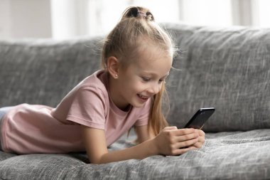 Gülümseyen küçük kız akıllı telefon kullanarak kanepede yatıyor.