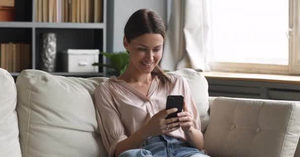 Giovane donna attraente sorridente che utilizza l'app per smartphone sul divano — Video Stock