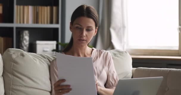 Jonge vrouw met papieren met laptop die rekeningen thuis betaalt — Stockvideo