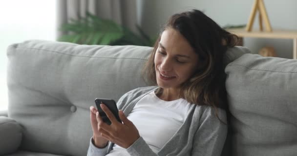 Šťastná mladá žena relaxuje na útulném gauči, pomocí smartphonu. — Stock video