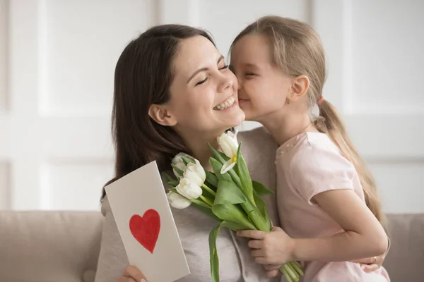 Liten dotter kysser mor på kinden, presenterar blommor och gåva — Stockfoto