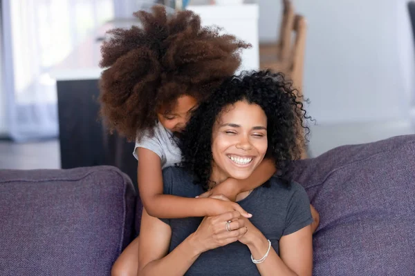 Little black girl hug smiling mom on couch from behind