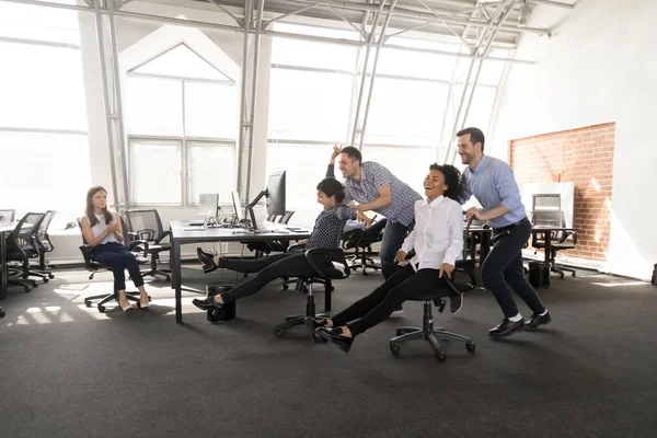 Excited diverse workers have fun racing on chairs in office