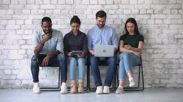 Etnia diversa jovens sentados em cadeiras usando dispositivos tecnológicos — Vídeo de Stock