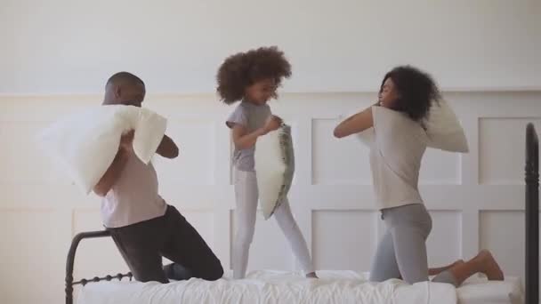 Alegría pequeña niña africana étnica luchando almohadas con los padres . — Vídeos de Stock