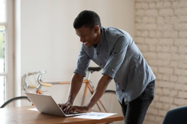 Gülümseyen Afrikalı Amerikalı iş adamı dizüstü bilgisayar kullanıyor, masanın yanında duruyor.