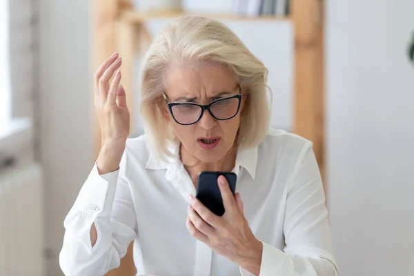 Mature businesswoman holding smartphone feels indignant having wi-fi connection problem — ストック写真