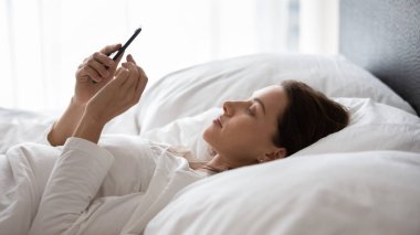 Genç bir kadın telefon kullanıyor, yatakta uzanıyor, sabahları sohbet ediyor.