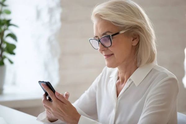 Focused middle-aged businesswoman seated at workplace using smart phone — ストック写真