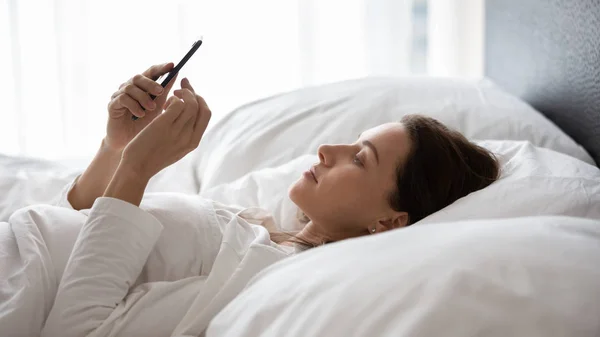 Mujer joven usando el teléfono, acostada en la cama, charlando por la mañana — Foto de Stock