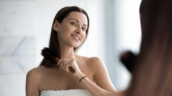 Sonriente hermosa mujer cepillando el cabello, mirando en el espejo en el baño —  Fotos de Stock