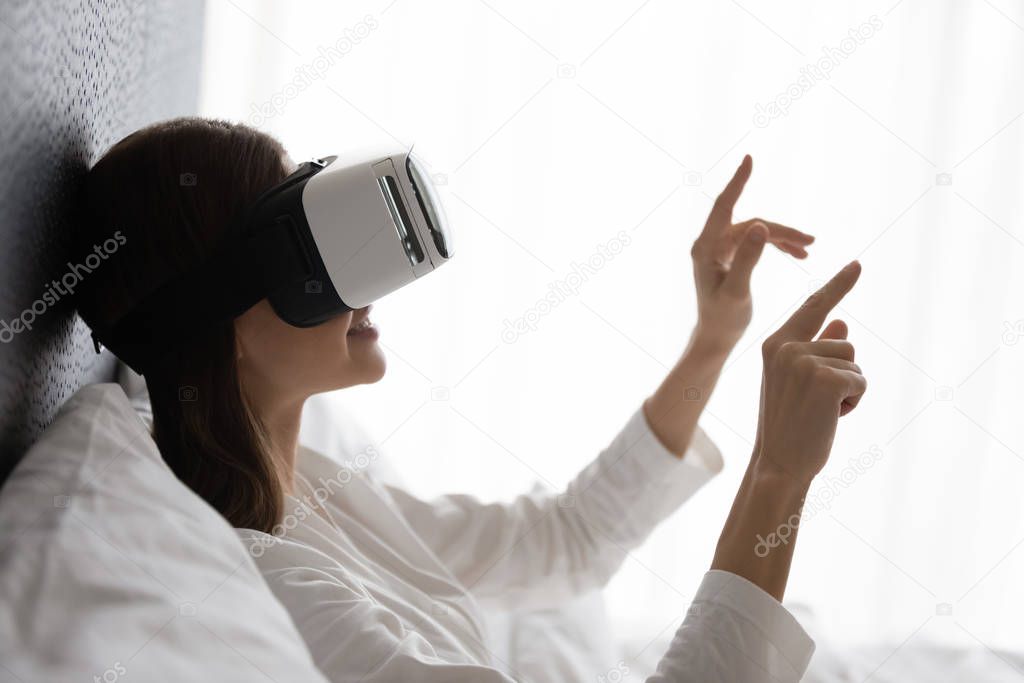Smiling woman wearing vr glasses sitting in bed, touching air