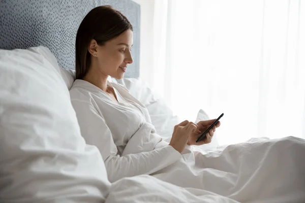 Mujer joven sonriente usando el teléfono, sentada debajo de una manta en la cama — Foto de Stock