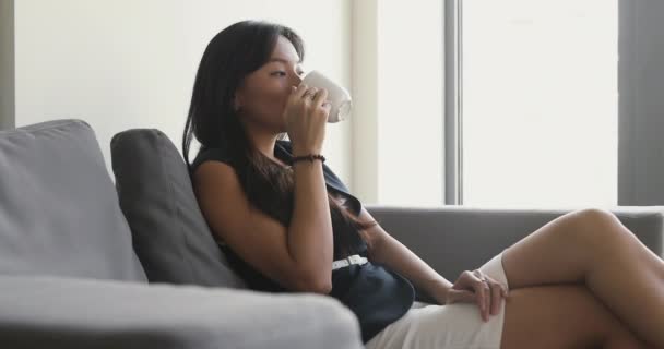 若いアジアのビジネスマンの女性はコーヒーを飲みながらソファでリラックス. — ストック動画