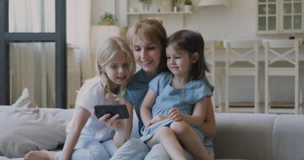 Счастливая женщина обнимает маленьких девочек, показывает смешные видео на смартфоне . — стоковое видео