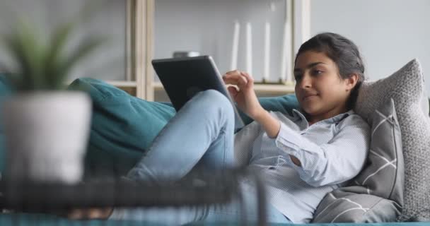 Ευτυχισμένη Ινδή γυναίκα χαλαρώνοντας στον καναπέ κρατώντας χρησιμοποιώντας ψηφιακό δισκίο — Αρχείο Βίντεο