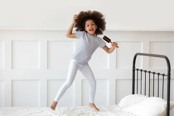 Chica saltando en la cama sosteniendo cepillo de pelo como micrófono y cantando — Foto de Stock