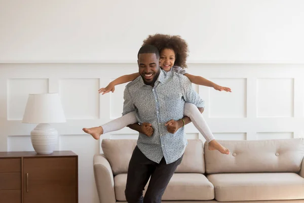 African father piggybacking little daughter playing together in living room