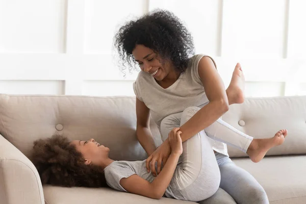 African mother tickles daughter sitting on couch have fun indoors — Stock Photo, Image