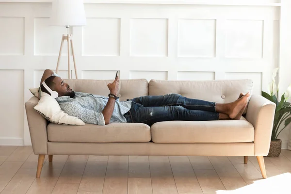 Afrikansk mann med hodetelefoner som hviler på sofaen med smarttelefon – stockfoto