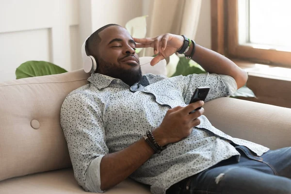 Africano cara segurando smartphone olhos fechados ouvir música em fones de ouvido — Fotografia de Stock